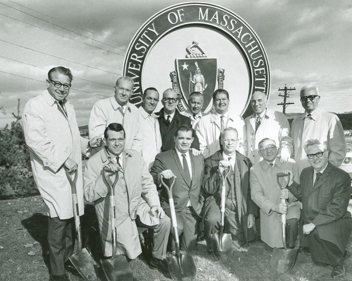 old ground breaking photo, 13 men holding shovels, large University of Massachusetts Medical School logo/structure behind them 